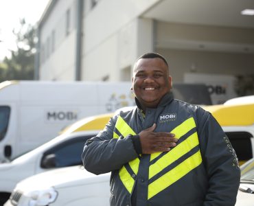 Funcionário da Mobi uniformizado com a jaqueta da empresa, sorrindo e batendo no peito. Ao fundo, parte da frota de veículos da Mobi