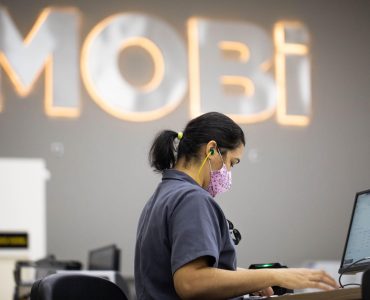 Funcionária da Mobi sentada em frente a um computador, utilizando máscara e protetor auricular. Ao fundo, o logo da Mobi inserido em uma parede cinza.