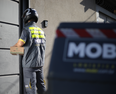 Motofretista da Mobi segurando uma caixa para realizar uma entrega. Em frente, está o baú de transporte da moto, com logo da Mobi.