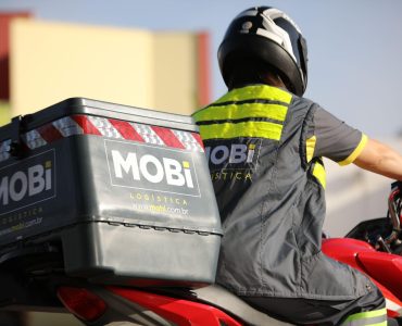Motofretista da Mobi de costas, usando uma jaqueta da Mobi, em cima da moto com baú de transporte.