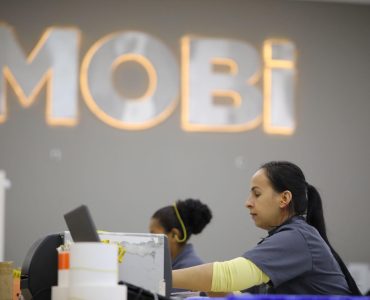 Colaboradora da Mobi Logística trabalhando na triagem de remessas. Ao fundo, desfocado, o logo da Mobi.