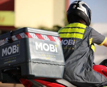  Motofretista conduzindo uma moto vermelha, Em destaque o baú da moto com o logo da Mobi Logística.