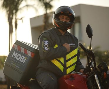 Motofretista da Mobi uniformizado, em cima de uma moto vermelha que está parada. O motofretista bate no peito, próximo ao logo da marca estampado na jaqueta.