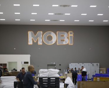 Galpão da Mobi com alguns funcionários realizando o serviço de separação de remessas. Ao fundo, na parede, o logo da Mobi.