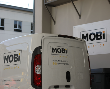 Carro da Mobi Logística parado na saída de uma garagem, com logo da Mobi ao fundo na parede.