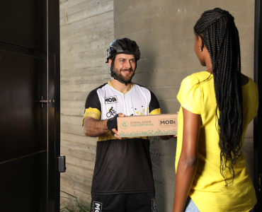 Entregador ciclista da Mobi Logística entregando uma embalagem sustentável para uma mulher, na porta de casa.