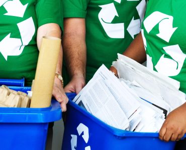 Pessoas com camiseta verde de símbolo de reciclagem, segurando caixas azuis com papéis.