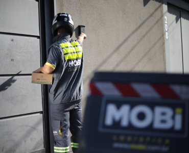 Motofretista da Mobi Logística tocando o interfone de uma casa, segurando uma caixa da Mobi embaixo do braço.