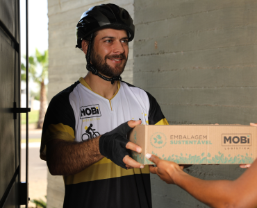Entregador ciclista da Mobi Logística, entregando uma caixa de embalagem sustentável a uma pessoa, na porta da casa dela.