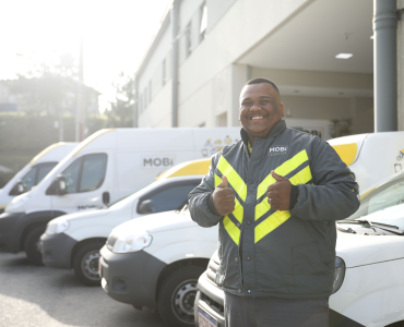 Funcionário da Mobi Logística fazendo sinal de positivo com as mãos, com os carros de entrega da Mobi ao fundo.