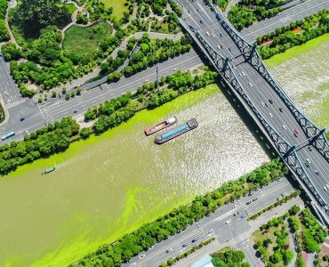 Imagem aérea de um rio que passa no meio de uma cidade, com uma ponte cruzando ele e navios de carga navegando por ele.