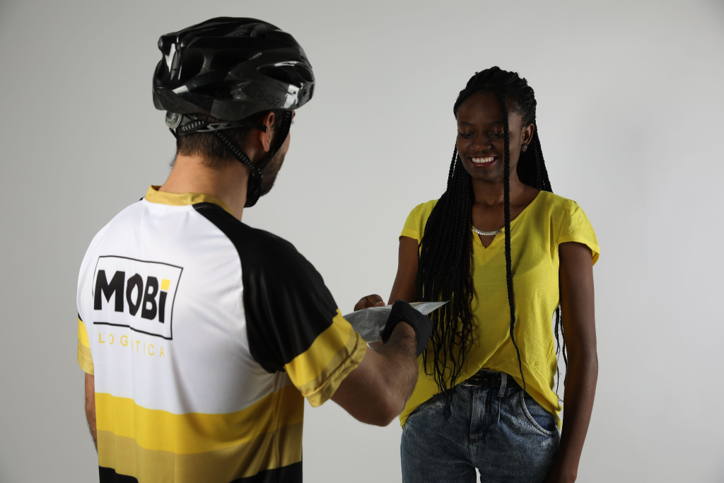 Mulher de amarelo recebendo pacote do entregador ciclista da Mobi Logística.