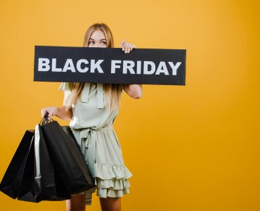 Mulher segurando uma placa com a frase “Black Friday” em frente ao rosto com uma mão; com a outra ela segura sacolas pretas de compra.