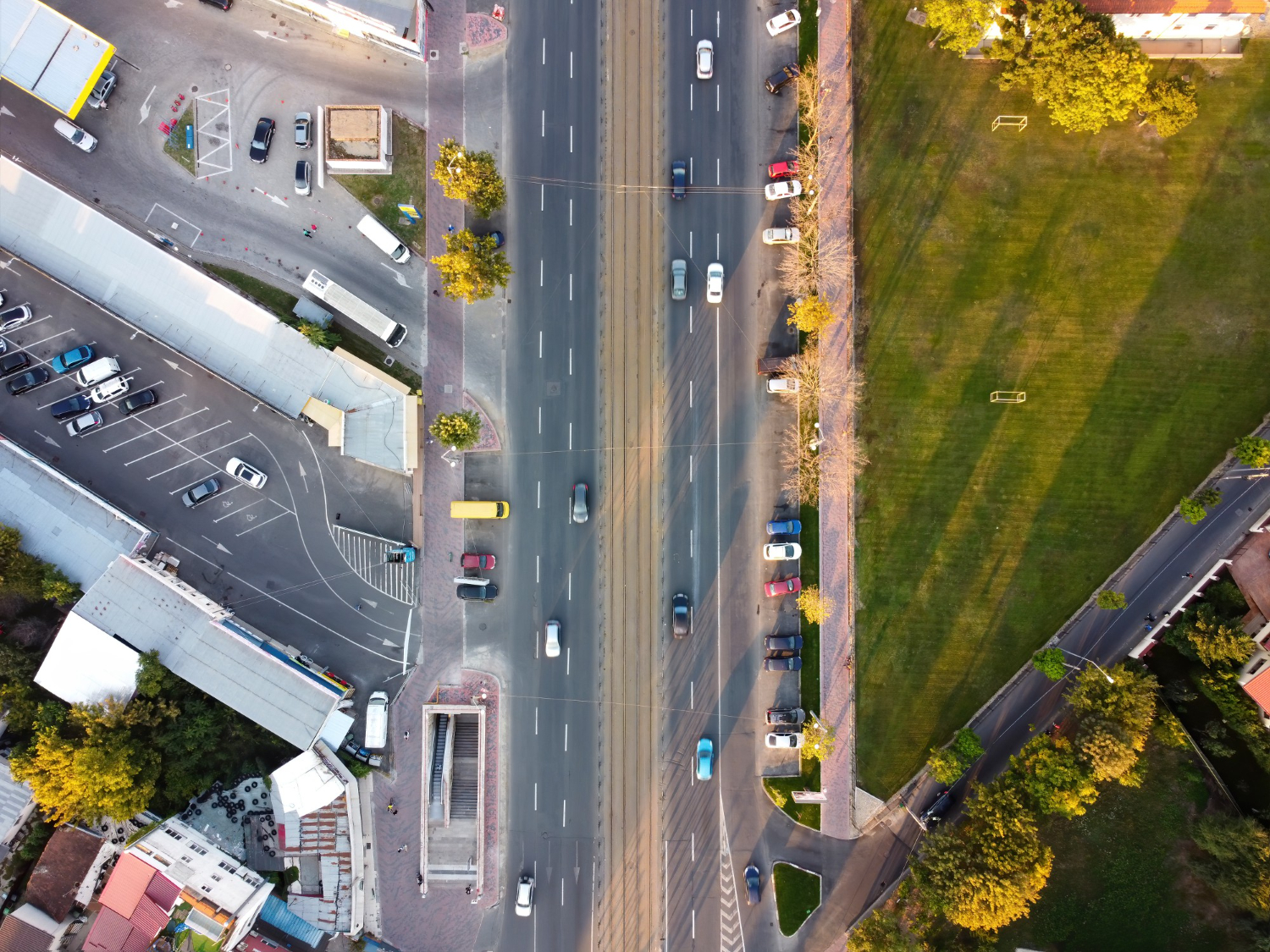 Vista superior de duas ruas com alguns carros em movimento. Ao lado de uma das ruas, um estacionamento; do outro, uma área verde